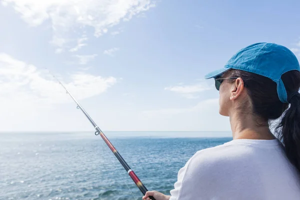 Sahilde balık tutan bir kadın. Güneş gözlüğü ve şapka takıyor..