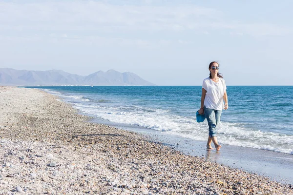 Kumsalda yürüyüş yapan bir kadın. Sıradan kıyafetler giyiyor ve sandaletlerini elinde tutuyor..