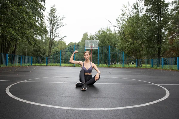 Fitness model in a sports top and leggings doing sports exercises on sports grounds in the park. The model shows the muscles