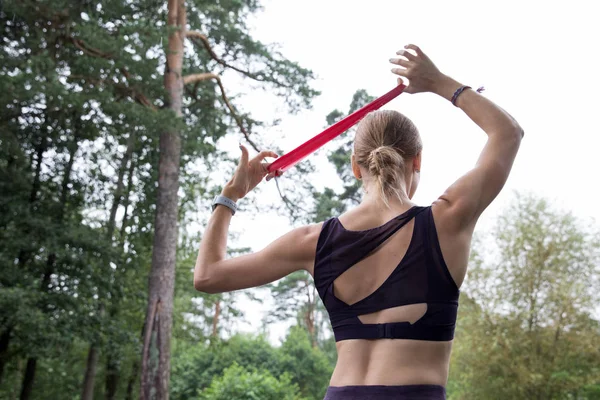 Beautiful girl in sports top and leggings trains with red elastic rope on sports grounds in the park.Training in the rain