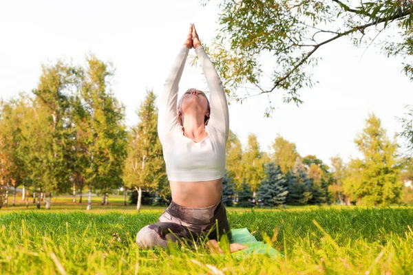 A girl in a white t-shirt and sports leggings doing yoga in the park