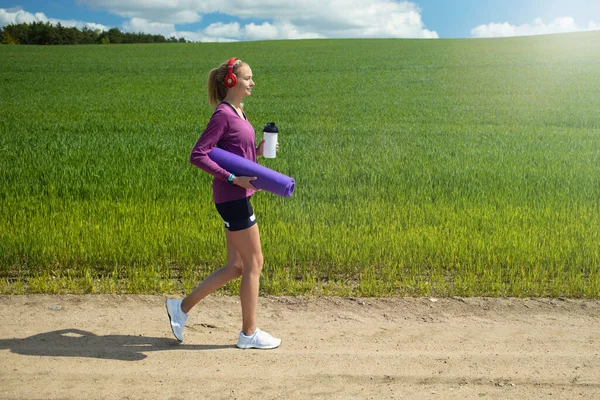 女の子は水のボトルと緑のフィールドに対してロールアップフィットネスマットを保持しています 新鮮な空気の中で夏のトレーニング — ストック写真