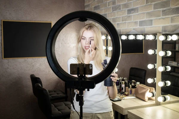 A girl stands in front of a ring lamp and conducts online courses on applying makeup in a beauty studio. Using makeup brushes