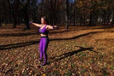 Parlak eşofmanlı bir kız parkta dambıllarla egzersiz yapıyor.