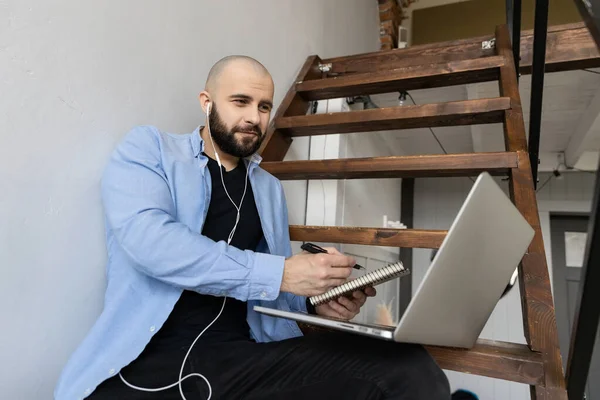 A guy in a blue shirt and black jeans is sitting on the stairs in the house and working at the computer. The concept of freelance work