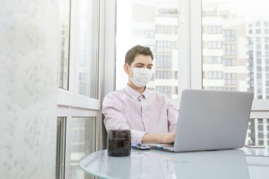 Koruyucu yüz maskeli bir adam virüs salgını sırasında evden bilgisayarın başında çalışıyor. Virüs enfeksiyonu sırasında karantina