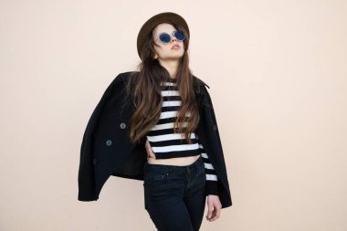 Paltolu, gözlüklü ve şapkalı güzel bir esmerin portresi. Sokakta duvara karşı poz veriyor.