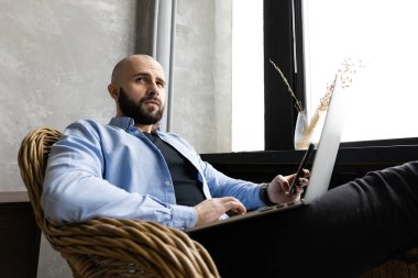 Mavi gömlekli adam evde, pencerenin yanında bir dizüstü bilgisayar üzerinde çalışıyor. Uzaktan çalışma kavramı.