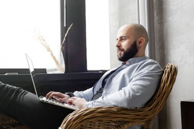 Mavi gömlekli adam evde, pencerenin yanında bir dizüstü bilgisayar üzerinde çalışıyor. Uzaktan çalışma kavramı.