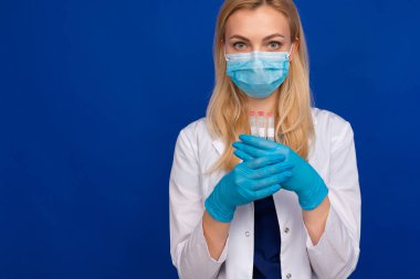 Tıbbi maskeli ve eldivenli bir kız doktor mavi arka planda test tüpleri tutuyor.