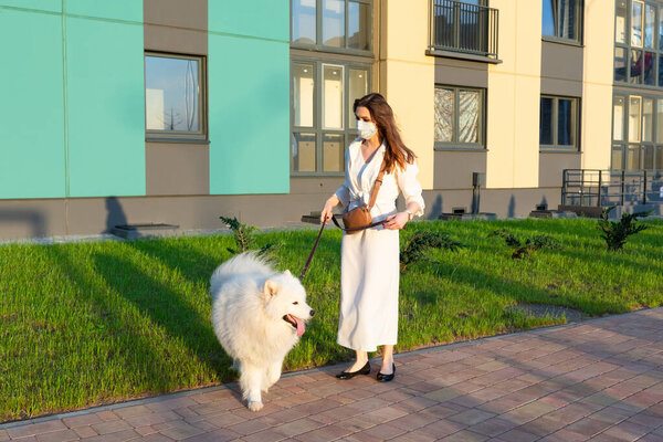 Девушка в платье и защитная маска на лице гуляет с белой пушистой собакой в жилом районе