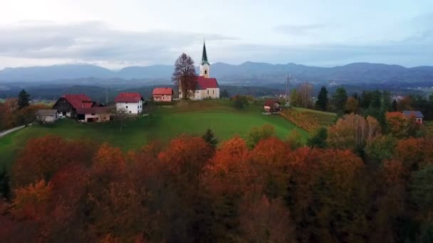 航空写真 スロベニア語の田舎で秋の早朝の日の出に飛んでいます 丘の上に小さな教会に向かって美しい森に行く — ストック動画