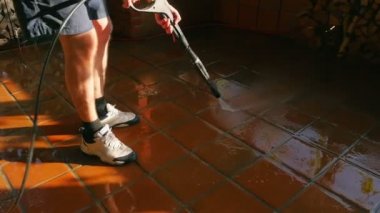 Erkek şort yüksek basınçlı yıkayıcı ile açık hava teras karoları temizler. Su serpme ve akşam günbatımında parlayan. Temizlik civarı ev bahar.