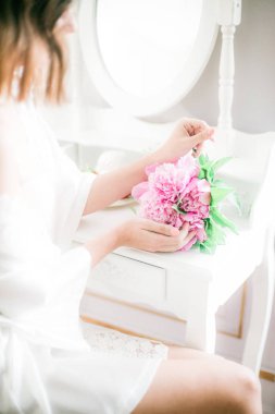  Çok güzel bir gelin bir peonies beyaz bir tuvalet masası ayna, inci kolye ile oturup onu elinde tutuyor. Gelin sabah hazırlık.