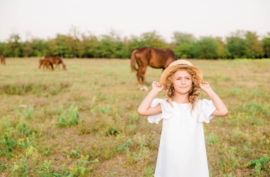 Beyaz bir elbise ve hasır şapka alanındaki atları ile hafif kıvırcık saçlı tatlı bir küçük kız. Kırsal yaşam Güz. Atlar ve insanlar