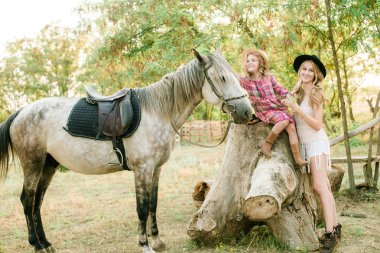 Beautiful young girl with blond hair in a suede jacket with fringe with little sister in a straw hat and checkered vintage dress with a horse in the countryside on a sunny autumn day clipart