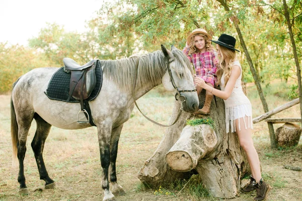 美丽的年轻女孩与金发在一个麂皮绒夹克与小妹妹在一个稻草帽和格子复古礼服与马在阳光明媚的秋天的一天在农村 — 图库照片