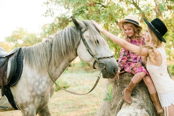 晴れた秋の日に田舎で馬と麦わら帽子と市松模様のヴィンテージのドレスのリトル シスターをフリンジ付きスエード ジャケットのブロンドの髪を持つ美しい少女 — ストック写真