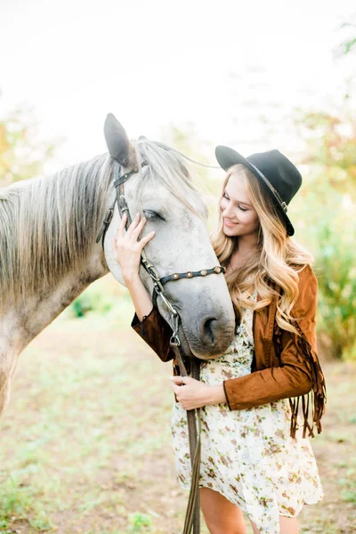 美丽的年轻女孩 金发碧眼 披着条纹的麂皮夹克 戴着黑色的软盘帽 在日落时微笑着抚摸着她的马 — 图库照片