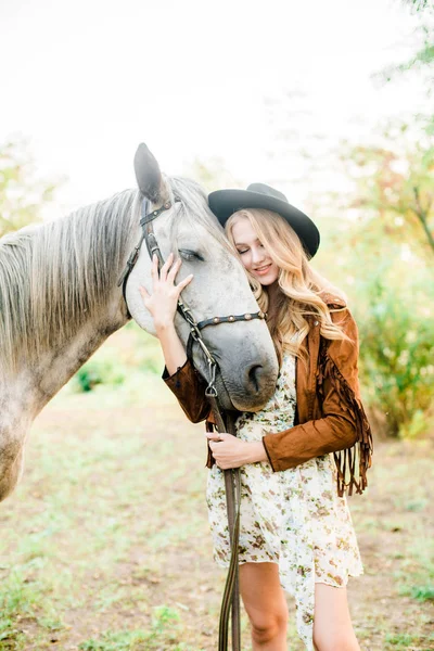 美丽的年轻女孩 金发碧眼 披着条纹的麂皮夹克 戴着黑色的软盘帽 在日落时微笑着抚摸着她的马 — 图库照片
