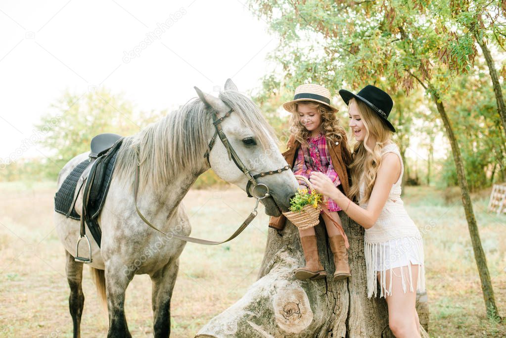 Beautiful young girl with blond hair in a suede jacket with fringe with little sister in a straw hat and checkered vintage dress with a horse in the countryside on a sunny autumn day