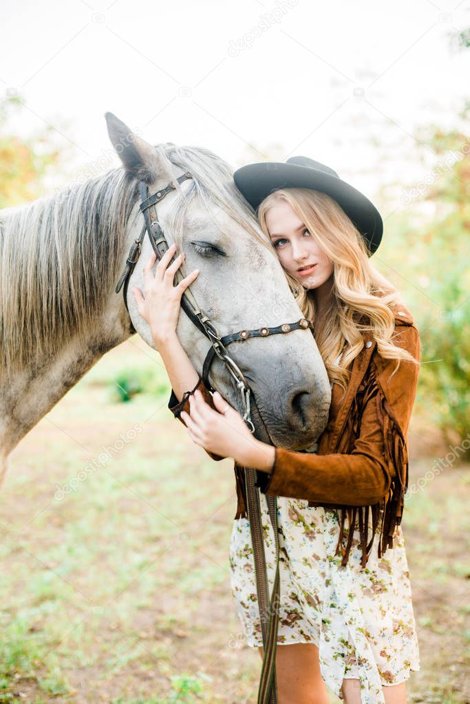 Beautiful  young girl with blond hair in a suede jacket with a fringe , wearing black floppy hat, smiling and stroking her horse in sunset