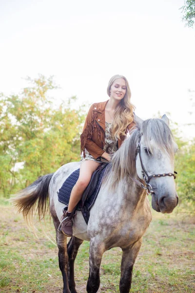 美丽的年轻女孩 长着金色的头发在一个麂皮绒夹克与条纹微笑 骑着马在秋季日落 — 图库照片