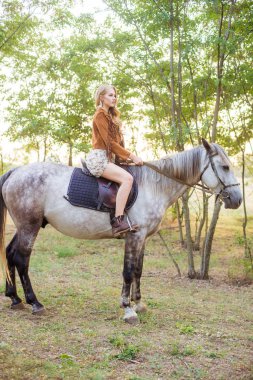 Beautiful  young girl with long blond hair in a suede jacket with a fringe smiling and astride a horse in sunset in autumn clipart
