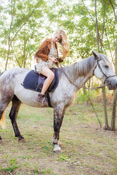 Beautiful  young girl with long blond hair in a suede jacket with a fringe smiling and astride a horse in sunset in autumn