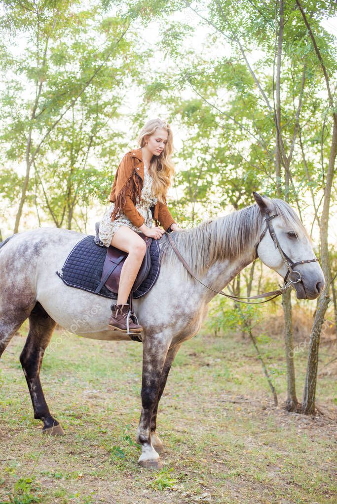 Beautiful  young girl with long blond hair in a suede jacket with a fringe smiling and astride a horse in sunset in autumn