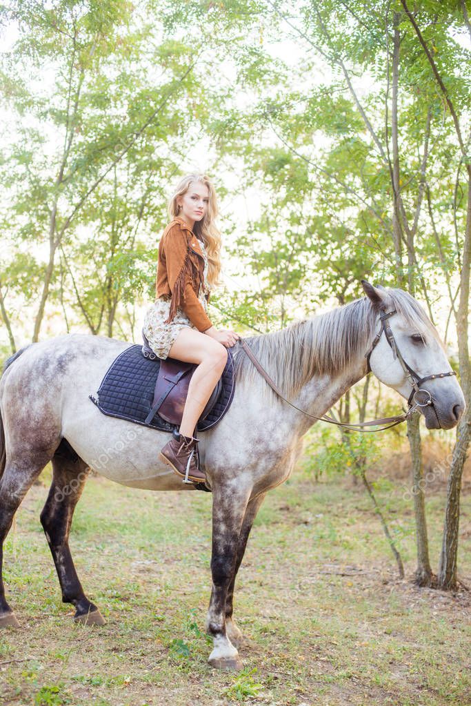 Beautiful  young girl with long blond hair in a suede jacket with a fringe smiling and astride a horse in sunset in autumn