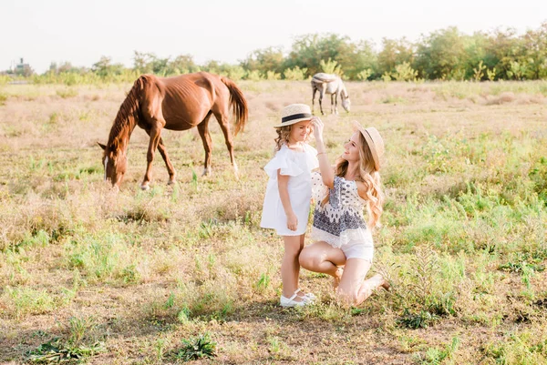 一个美丽的年轻女孩 在一顶草帽与一个小妹妹拥抱和笑附近的马 在农村 温暖的秋天在一个阳光明媚的日子 与轻卷曲的头发 — 图库照片