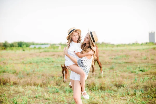 一个美丽的年轻女孩 在一顶草帽与一个小妹妹拥抱和笑附近的马 在农村 温暖的秋天在一个阳光明媚的日子 与轻卷曲的头发 — 图库照片