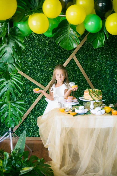 A festive candy bar for a little girl, decorated in a tropical style with a lemon cake cupcakes and marshmallows and bright balloons. Children\'s party in a tropical style with yellow, green and black balloons and tropical leaves