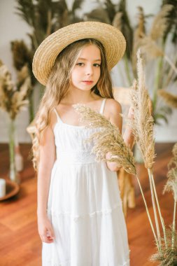 Uzun sarı saçlı beyaz bir sarafan ve bir hasır şapka rattan koltuk ve reed dekor ile tatlı bir küçük kız