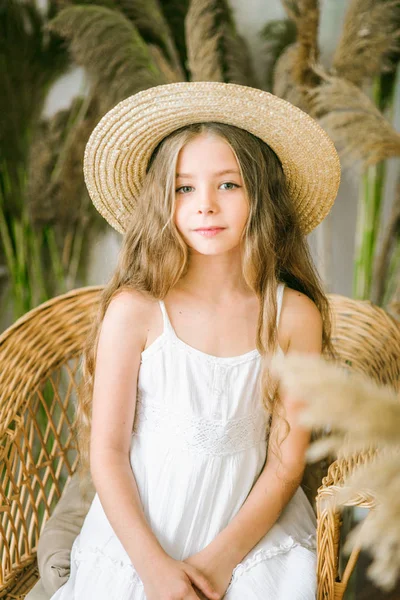 一个可爱的小女孩 长着金色的头发 在白色的 Sarafan 和一顶草帽在藤椅和芦苇装饰 — 图库照片
