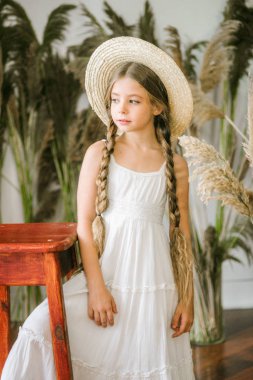 Uzun sarı saçlı beyaz bir sarafan ve bir hasır şapka rattan koltuk ve reed dekor ile tatlı bir küçük kız