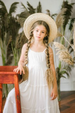 Uzun sarı saçlı beyaz bir sarafan ve bir hasır şapka rattan koltuk ve reed dekor ile tatlı bir küçük kız