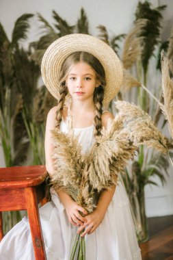 Uzun sarı saçlı beyaz bir sarafan ve bir hasır şapka rattan koltuk ve reed dekor ile tatlı bir küçük kız