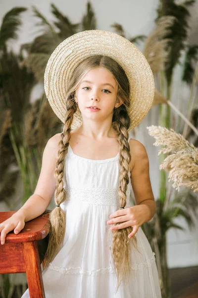 Uzun sarı saçlı beyaz bir sarafan ve bir hasır şapka rattan koltuk ve reed dekor ile tatlı bir küçük kız
