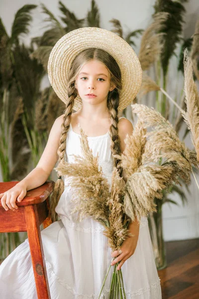 一个可爱的小女孩 长着金色的头发 在白色的 Sarafan 和一顶草帽在藤椅和芦苇装饰 — 图库照片