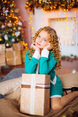 Kıvırcık saçlı sarışın evde hediyeler ve çelenk ve dekore edilmiş bir şömine ile Noel ağacı yakınındaki sevimli küçük kız 