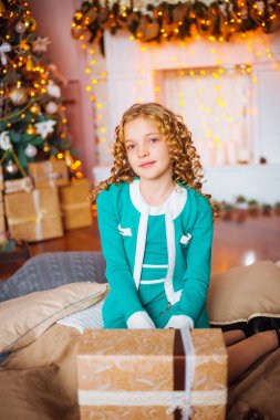 Kıvırcık saçlı sarışın evde hediyeler ve çelenk ve dekore edilmiş bir şömine ile Noel ağacı yakınındaki sevimli küçük kız 