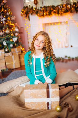Kıvırcık saçlı sarışın evde hediyeler ve çelenk ve dekore edilmiş bir şömine ile Noel ağacı yakınındaki sevimli küçük kız 