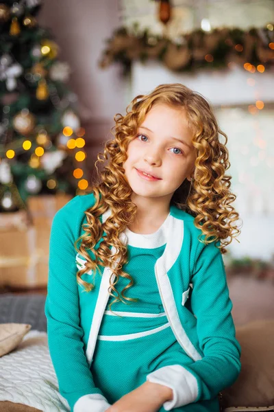 金髪巻き毛自宅ギフトと花輪と枕の上に座って装飾が施された暖炉とクリスマス ツリーの近くでかわいい女の子 — ストック写真