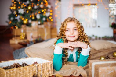 Kıvırcık saçlı sarışın evde hediyeler ve çelenk ve dekore edilmiş bir şömine ile Noel ağacı yakınındaki sevimli küçük kız 
