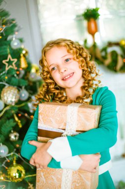 Kıvırcık saçlı sarışın evde hediyeler ve çelenk ve ekoseler ve yastıklar üzerinde oturan bir şekilde dekore edilmiş bir şömine ile Noel ağacı yakınındaki sevimli küçük kız