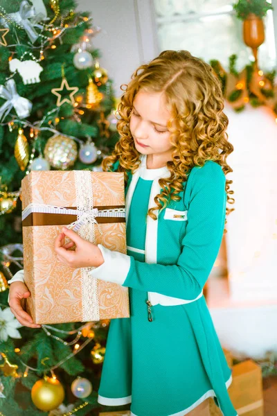 Kıvırcık saçlı sarışın evde hediyeler ve çelenk ve ekoseler ve yastıklar üzerinde oturan bir şekilde dekore edilmiş bir şömine ile Noel ağacı yakınındaki sevimli küçük kız