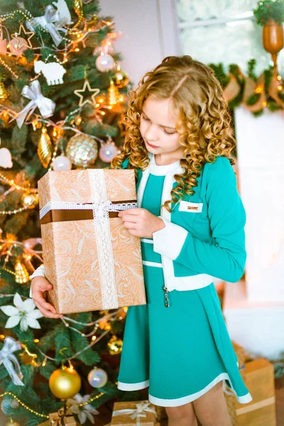 Kıvırcık saçlı sarışın evde hediyeler ve çelenk ve ekoseler ve yastıklar üzerinde oturan bir şekilde dekore edilmiş bir şömine ile Noel ağacı yakınındaki sevimli küçük kız