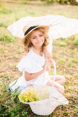 Işık ile tatlı bir küçük kız bir hasır şapka, beyaz bir elbise ve dantel şemsiye yaz tarlada çiçekler bir sepet ile bukleler. Yaz aylarında kırsal yaşam